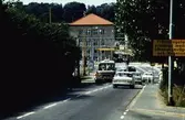 Vy från Kvarnbygatan mot uppresta järnvägsbommar där en buss och bilar passerar, 1970-tal. I bakgrunden ses Hantverkshuset (Kungsbackavägen 2/Frölundagatan 1) i Mölndals Bro.