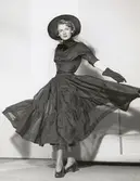 Nordiska Kompaniet. Kvinna i svart festklänning med trekvartsärm. Snävtliv med hög knäppning och rosett under hakan. Vid kjol med vågräta rynkade våder. Bred svart hatt med flor i nacken. text på baksidan: 