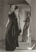 Nordiska Kompaniet. Kvinna i mörk aftonklänning av sammet framför en spegel. Turnyrliknande drapering i sidan. Text på baksidan: 
