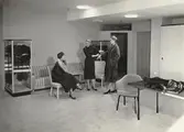 Nordiska Kompaniet, interiör. Modell visar upp en päls för en sittande kund. Text på baksidan: