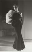 Nordiska Kompaniet. Kvinna i mörk, snäv aftonklänning, utsvängd från knäna. Ljus pälsboa/muff över ena axeln. Text på baksidan: nov 1950