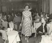 Nordiska Kompaniet. Tonårsmode 1950. Interiör med flicka i diagonalmönstrad prickig klänning med v-ringning och liten holkärm. Vitt skärp i midjan. I bakgrunden modepublik vid dukade kaffebord. Text på baksidan:
