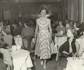 Nordiska Kompaniet. Tonårsmode 1950. Interiör med flicka i diagonalmönstrad prickig klänning med v-ringning och liten holkärm. Vitt skärp i midjan. I bakgrunden modepublik vid dukade kaffebord. Text på baksidan: