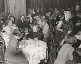 Nordiska Kompaniet. Tonårsmode 1950. Kvinna i mörk rutig klänning med kort ärm. Runt henne en ung modepublik vid dukade bord med läskedrycksflaskor. Text på baksidan: 
