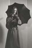 Nordiska Kompaniet. Kvinna i mörk kappa med kapuschong och uppfällt paraply. Text på baksidan: 