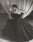 NK:s Franska damskrädderi. Kvinna i aftonklänning av mörk tyll. Kring ringningen arr arrangemang med fjädrar. Text på baksidan: 