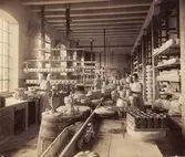 Rörstrands fabrik. Fabriksinteriör, verkstadshuset. Kvinnor och män i arbete vid bänkar. Porslin i långa rader längs bord och hyllor. 