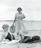 Kvinna i klänning, kort jacka och stråhatt i handen står på en strand. Två kvinnor i baddäkt ligger ned på stranden.