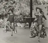 Två kvinnor från Operabaletten i bodys, hattar och långa, blommiga kappor dansar på scen i Kungsträdgården i Stockholm, för Nordiska Kompaniet.