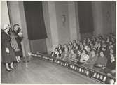 Tonårsmodevisning på Borgarskolan. Kvinna i kavaj, kjol och hatt på scen, bredvid kvinna som pratar i mikrofon.