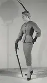 Modell i dräkt i brun melerad tweed, hatt med hög fjäderpenna, handskar och paraply. Från Jacques Fath.
