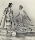 Kvinna till vänster i randig klänning och pumps, kvinna till höger i vit ärmlös blus och kjol med korg på armen, står på en solstol.