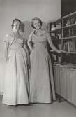 Tonårsmode. Modell till vänster i rosa aftonklänning och bolero i rips och kvinna till höger i aftonklänning i blå rips, vid en bokhylla.