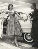 Tonårsmode. Modell i blommig klänning och man i kostym med cigarett i handen, står vid en bil.