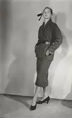 Modell i dräkt med skärp, hatt med dekorband och pumps. Från J. Griffe.