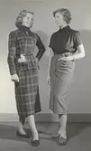 Tonårsmodevisning. Modell i rutig knytblus, kjol och skärp, bredvid modell i svart blus, pennkjol och skärp.