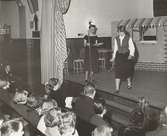 Tonårsmodevisning, Östra Real. Modell i väst, kjol och vit blus på scen bredvid kvinna med mikrofon. Publik i bänkrader.