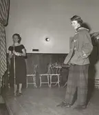 Tonårsmodevisning, Östra Real. Modell på scen i duffel och rutiga byxor. Kvinna pratar i mikrofon.