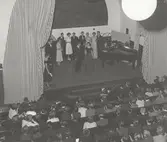 Tonårsmodevisning på Östra Real. Killar i kostym och tjejer i klänning förbereder dans på scen vid en flygel, framför publik.