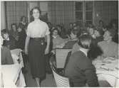 Tonårsmodevisning. Modell i blus, plisserad kjol, skärp med nitar och fickur hängande från skärpet. Publik sitter vid bord.