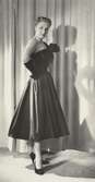 Modell i aftonklänning med rosett, handskar och pumps, framför draperi. Från Christian Dior.