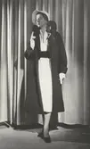 Modell i mörk kappa, vit klänning med brett skärp, vit hatt och vita handskar. Från J. Fath.