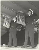 Två män i kostym och skärmhatt spelar trumpet med NK-loggan hängande från trumpeterna. Tonårsmode visas i Kungsträdgården.