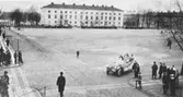 Torget med bil, som sannolikt deltager i biltävling. Med poliser och publik. Lennart Rydbergs samling