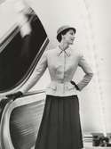 Modell i ljus jacka, veckad kjol, hatt och handskar vid rulltrappa. Original Patou.