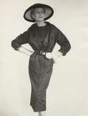 Modell i prickig klänning med skärp, stråhatt i lack, handskar och en garneringsblomma vid skärpet. Original Dior.