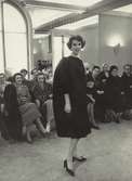 Modevisning på NK:s Franska damskrädderi, säsongens kollektion våren 1958. Mannekäng i mörk klänning, åskådare sitter på stolar..