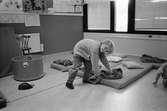 En pojke tittar leendes mot fotografen. Han lutar sig ned och håller på en filt som ligger på en madrass på golvet. På madrassen ligger en kudde. Kanske dags för en liten sovstund? Katrinebergs daghem, 1992.