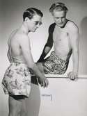 Två män i bar överkropp och badbyxor. Den ena badbyxan med mönster av surfande figurer och höga vågor. Nordiska Kompaniet. Från Nordiska Kompaniets kampanj med Californiakläder i maj 1947.