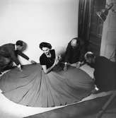 Modellen Hjördis Genberg fotograferas för Stil. Hon sitter på en vit matta, klädd i mörk överdel och vid kjol med en cigarett i handen. Tre män, fr. v. herr Paulsen, herr Hilgardt och kapten Ungervitter, draperar kjolen i en cirkel på golvet.
