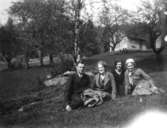 En okänd man, Clary Belfrage, Iris Persson (som gift) och Rosa Pettersson (f. Krantz) sitter i gräset framför en stuga, okänd plats och datum.