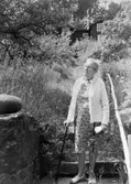 En äldre kvinna står nedanför trappan till Björkbo trädgård på Mogatan, 1970-tal.