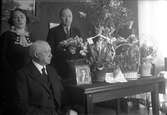 Gustav Anderssons far August fyller år och sitter vid ett bord med blommor och ett fotografi av sin fru Ottilia. Bakom honom står Gustav och hans syster Elsa.