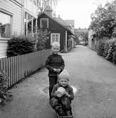På sparbanksgränden i Huskvarna leker två barn med en liten vagn. I husen på vänster sida ligger Gustavssons Tobak, Gamla Sparbanken, Arcadiahuset och Cykelhandlare W. Gustavssons eftr. G. Karlsson. Till höger finns Tvättetanterna.