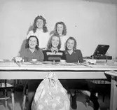 Fem flickor vid ett bord med en vevgrammofon i bakgrunden. Den ena flickan är klädd i vitt Lucialinne och har ljuskrona på huvudet. På bordet står kaffekoppar och små ljus och framför bordet en julklappssäck.