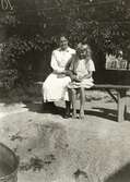 Nora Krantz (1879 - 1955) och dottern Rosa (1912 - 1994, gift Pettersson) sitter på en bänk utanför Stretered, cirka 1921 - 1923. Nora arbetade på Stretereds vårdhem, därav klädseln.