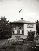 Nora Krantz (till vänster i vit hatt) och en okänd person sitter och fikar på Tulebokioskens veranda, 1930-tal. Utanför byggnaden står två träbänkar vid ett bord. På kioskens tak sitter en vimpel.