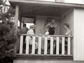Nora Krantz (till vänster i vit hatt) och en okänd ung bad-kille sitter och fikar på Tulebokioskens veranda, 1930-tal. En annan badklädd man häller upp kaffe till paret. Till vänster ses tall-grenar.