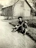 Rosa Krantz (1912 - 1994, gift Pettersson) och en liten pojke sitter vid vägkanten utefter Tulebovägen, 1930-tal. I bakgrunden ses vedboden som låg vid föräldrarnas personalbostad i Stretered.