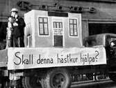 En lastbil passerar Rahms kappaffär på Östra Storgatan i Jönköping. På flaket är ett hus uppbyggt med två män bredvid. På dörren till huset finns texten: 