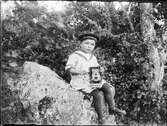 Liten pojke klädd i sjömanskostym sitter på en sten med en kamera i knät, Östhammar, Uppland