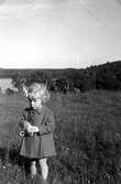 Eva Pettersson (född 1944, gift Kempe) står på en sommaräng, finklädd i kappa och med en blomma i håret år 1946. I bakgrunden ses Tulebosjön. Dotter till Bror och Rosa Pettersson.