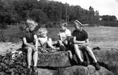 Eva Pettersson (född 1944, gift Kempe) sitter på en sten tillsammans med tre äldre pojkar (lekkamrater) vid Stenbryggan, Stretereds badplats, Tulebosjön år 1946. Eva är dotter till Rosa och Bror Pettersson.