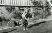 Karin Pettersson (född 1947) håller en vattenslang i handen på Gamlehagsvägen 17 i Torrekulla, cirka 1950. Bakom henne står hennes dockvagn utanför familjens villa.