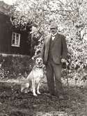 Trädgårdsmästare Johan Gustaf Johansson Scotte ståendes med en hund i en trädgård (troligtvis personalbostaden 