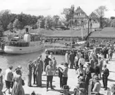Färjan Hebe V ligger i gamla hamnen på Visingsö. Mycket folk står på kajen och på stranden  väntar remmalag på att med häst och vagn få köra besökare på en guidad tur. Visingsborgs slottsruin syns i bakgrunden.